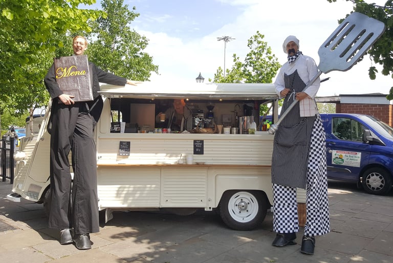 Performers on stilts dressed as a waiter and chef posing by a vintage mobile catering food truck.