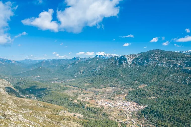 Panoramic view of a small mountain village nestled in a lush green valley under a bright blue sky.