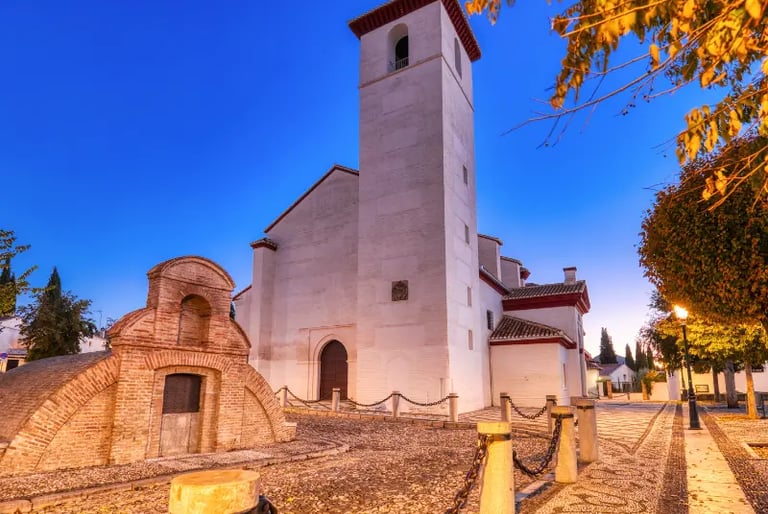 Historic white church and stone bell tower in a cobblestone plaza in Granada, Spain during twilight.