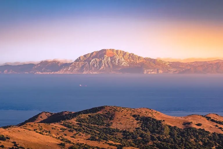 Scenic sunset view of the Rock of Gibraltar across the blue Mediterranean sea with coastal hills.