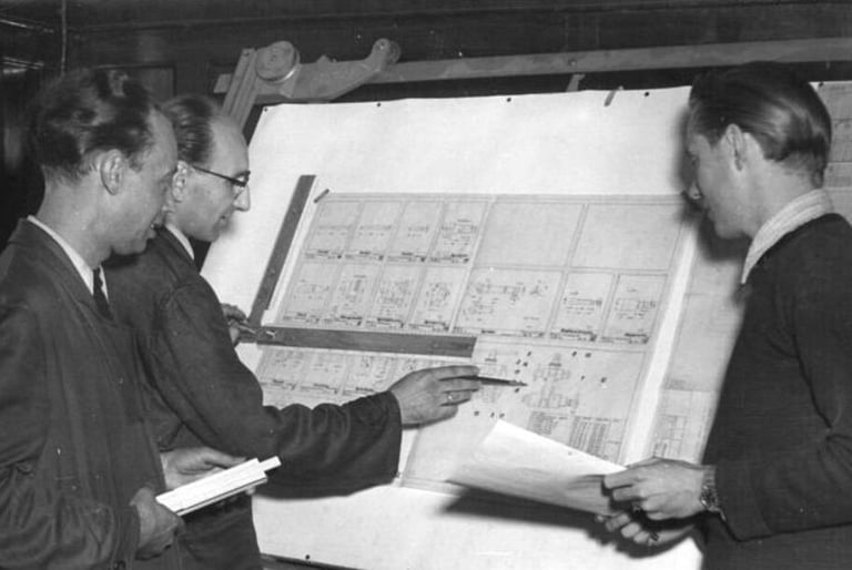 Vintage black and white photo of three engineers reviewing technical blueprints on a drafting board.