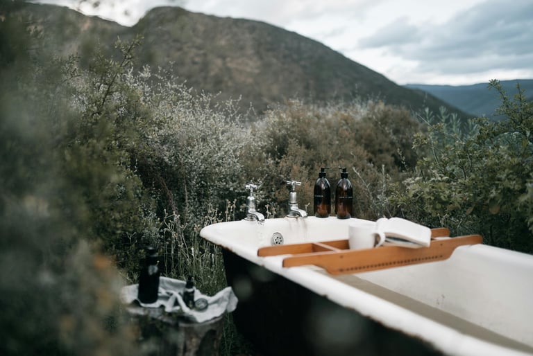 An outdoor clawfoot bathtub with a wooden tray and toiletries set against a scenic mountain landscape.