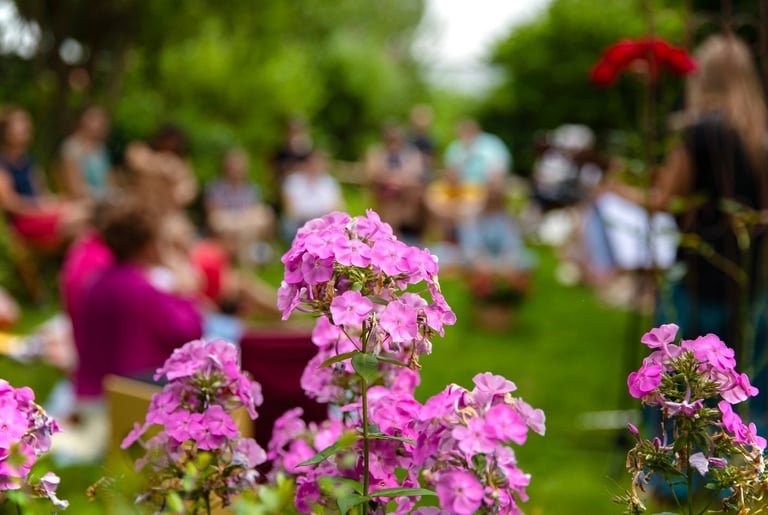 Im Vordergrund steht eine rosafarbene Blume. Im Hintergrund befindet sich eine Gruppe von Menschen, die im Garten zusammensit