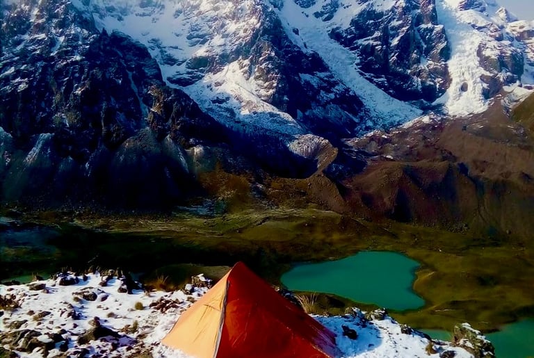 Orange tent at a snowy mountain campsite overlooking a turquoise alpine lake and glaciers in Peru. 