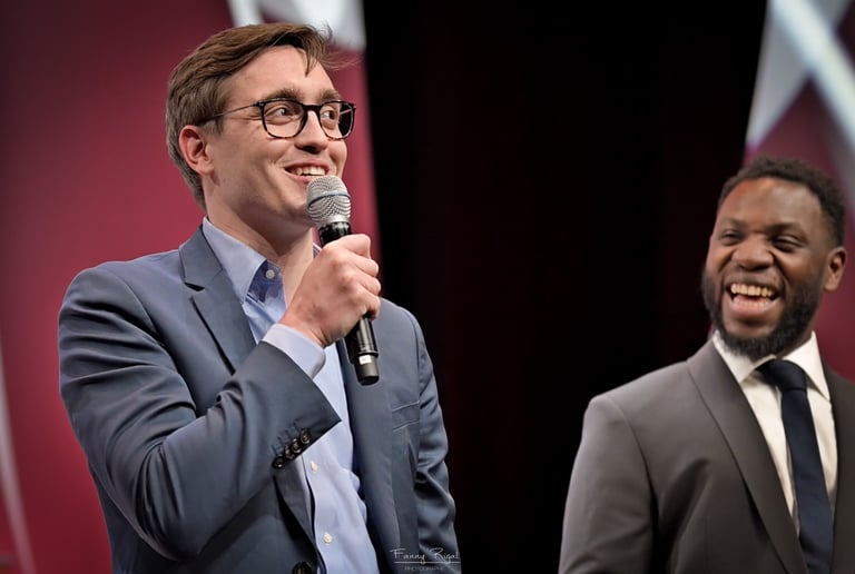 A smiling male speaker in a blue suit holding a microphone at a conference stage