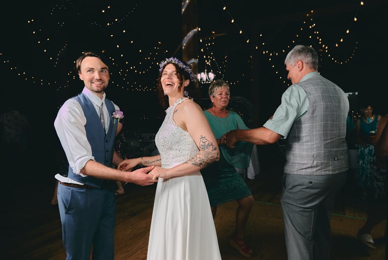 a bride and groom holding hands and dancing next to parents in a barn