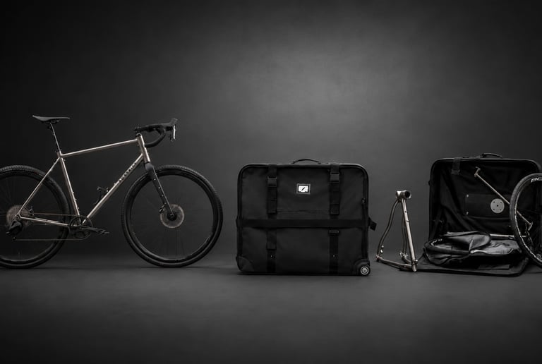 Titanium gravel bike next to a black padded bike travel bag and open storage case.