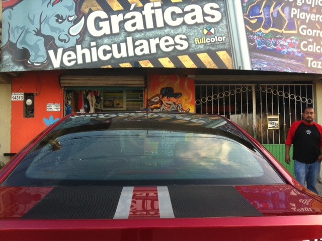 Red car with custom racing stripes parked in front of a vehicle graphics and wrap shop.