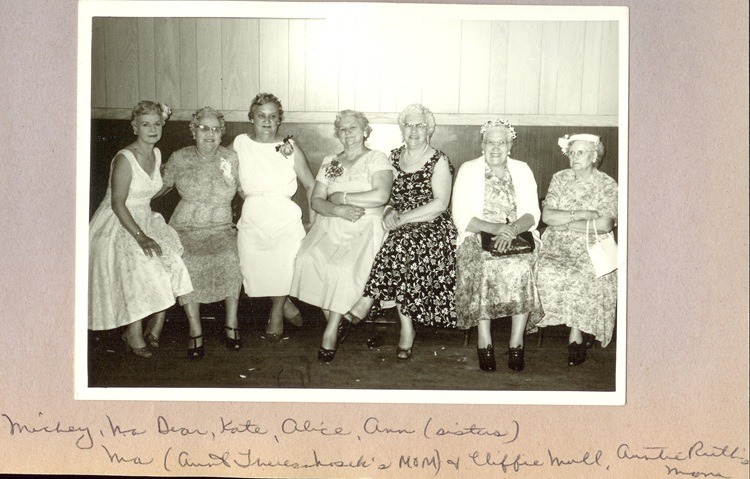 Vintage black and white photo of seven women in floral dresses sitting together at a formal gathering.