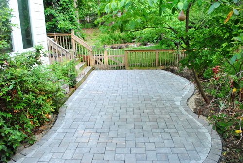 Curved grey stone paver patio installation next to a wooden deck and lush green backyard garden.