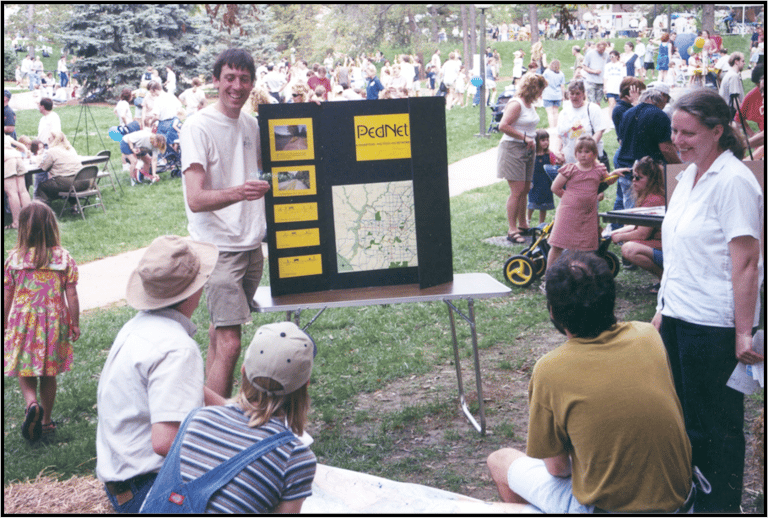 Older photo of a person presenting a trifold poster of info to a small crowd