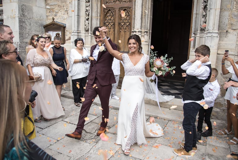Smiling bride and groom celebrate as guests throw flower petal confetti outside a stone church.