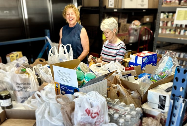 Two women standing behind table covered in bags of donated food in food pantry