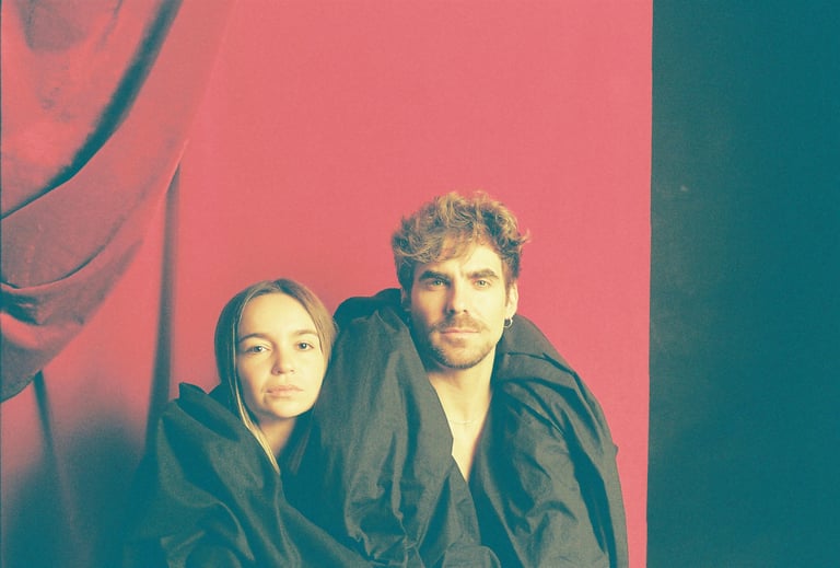 A man and woman posing wrapped in black fabric against a vibrant red backdrop.