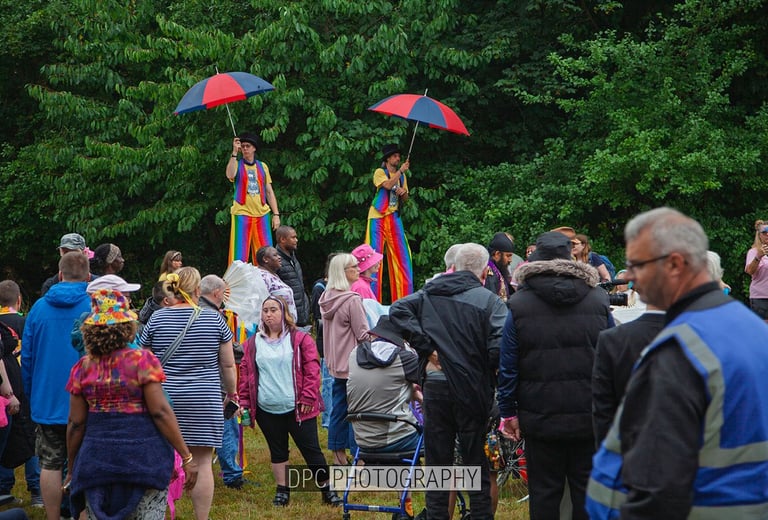 Performers on stilts with colorful umbrellas entertain a diverse crowd at an outdoor community festival.