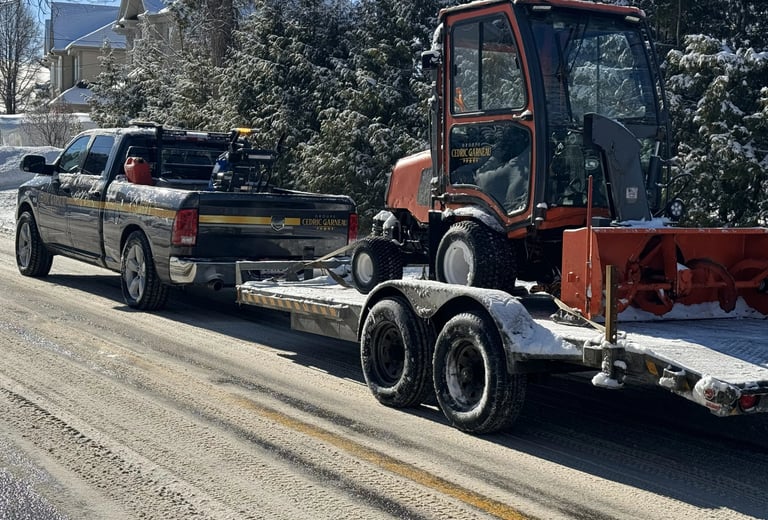 GCG pickup truck towing a trailer with a snow blower tractor on a snowy road.