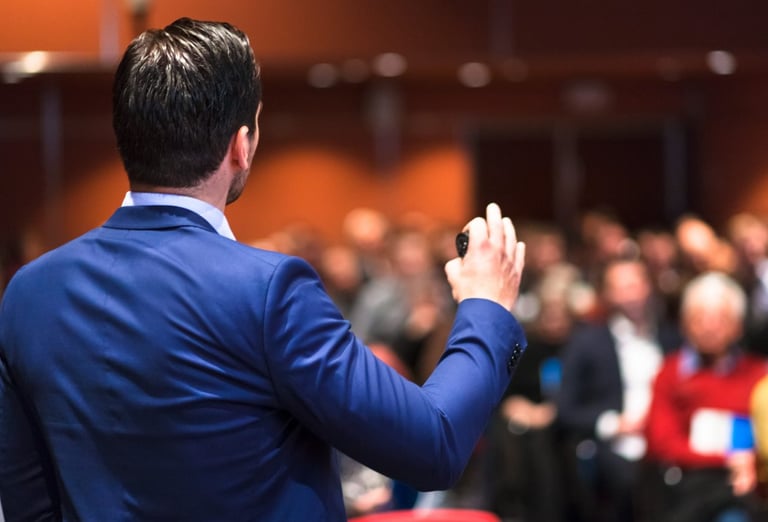 A keynote speaker in a suit presenting to a large audience at a conference.