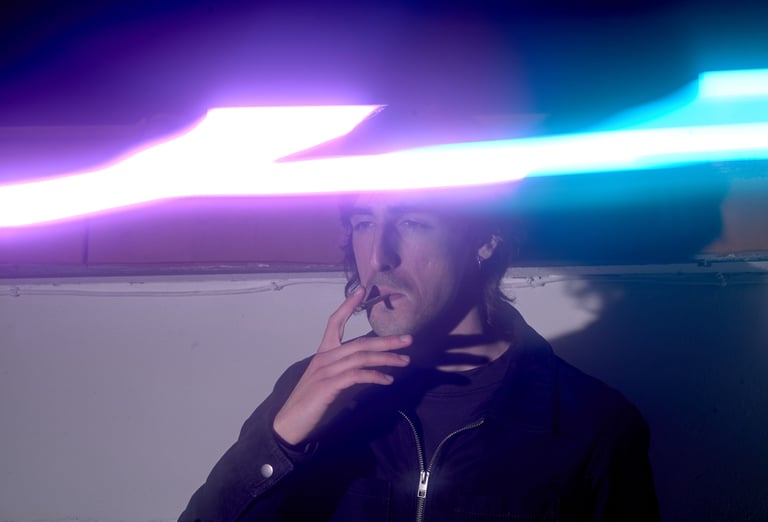 A man in a leather jacket smoking a cigarette under vibrant purple and blue neon light streaks.