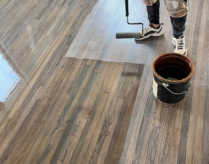 Professional applying a glossy polyurethane finish to dark stained oak hardwood floors using a paint roller.