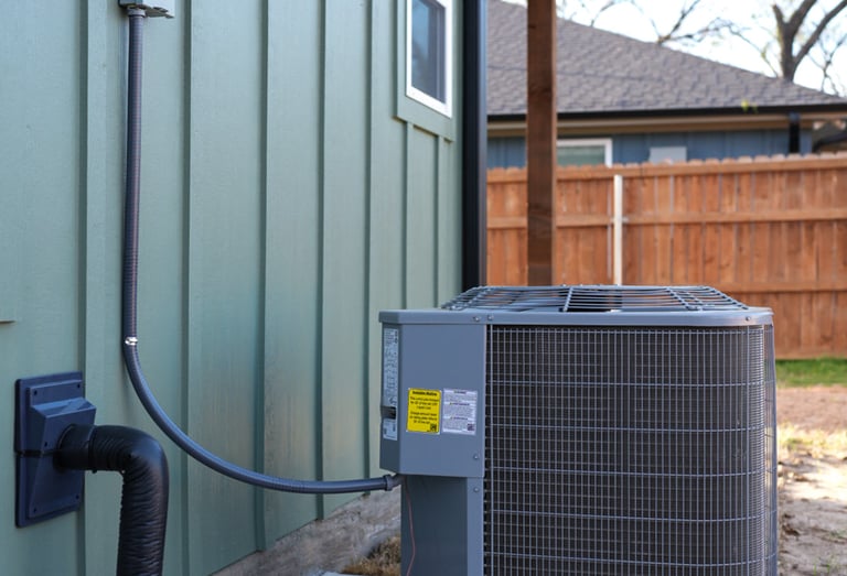 Exterior residential HVAC condenser unit installed on a concrete pad next to a green house wall.