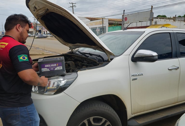 A mechanic installing a new Heliar car battery into a white Toyota Hilux truck with the hood open.