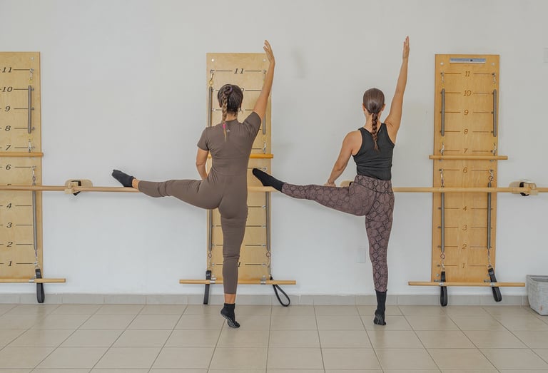 Two women performing a side leg raise stretch against the barre in a Pilates class.