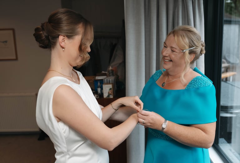 Smiling mother of the bride helps her daughter fasten a bracelet while getting ready for a wedding.