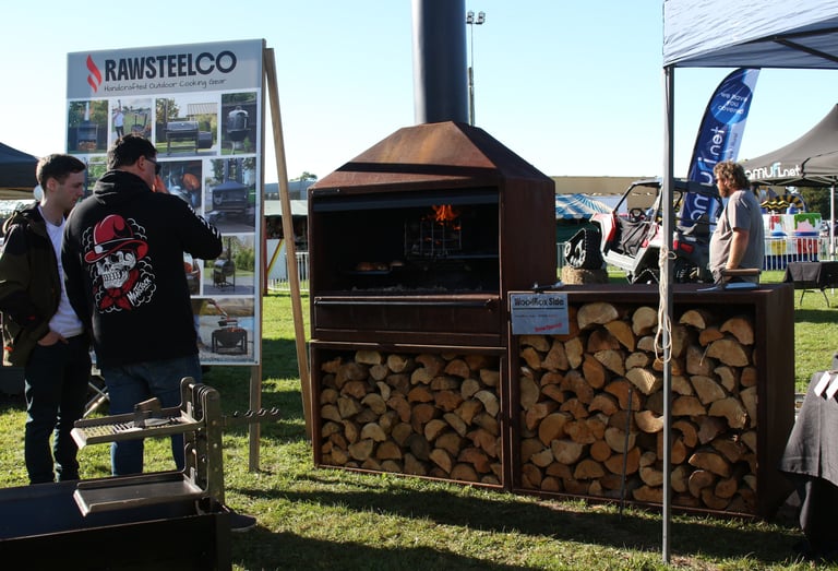 Customers at an outdoor fair viewing a RawSteelCo wood-fired oven with integrated firewood storage.