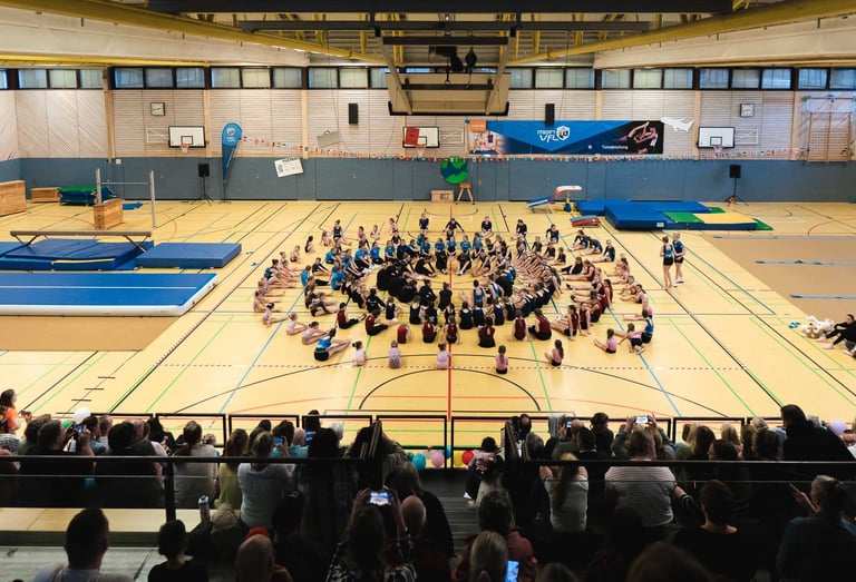 Viele Turnerinnen sitzen im Kreis auf dem Hallenboden vor Zuschauern. Eventfoto: Just Perspectives.