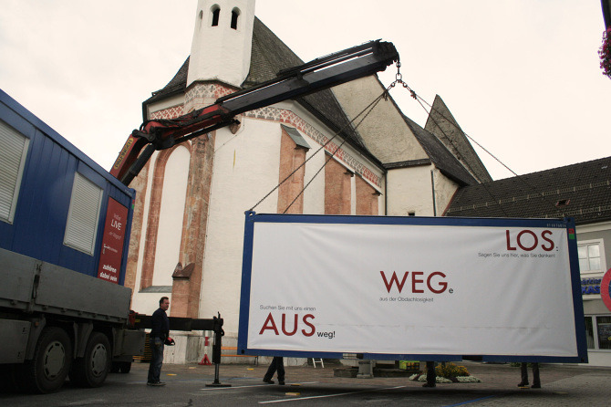 Kran bei der Aufstellung des Ausstellungscontainers "Aus weg los!" auf einem Dorfplatz.