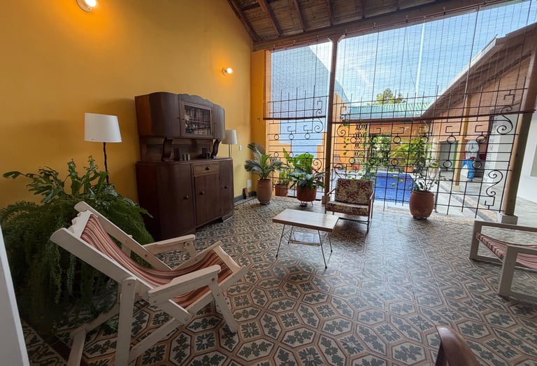 Rustic patio lounge with patterned floor tiles, antique wooden furniture, and a view of a garden swimming pool.
