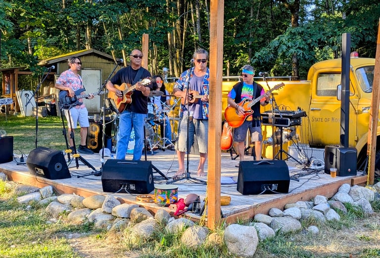 Live band performing outdoor music on a wooden stage with a yellow vintage truck background.