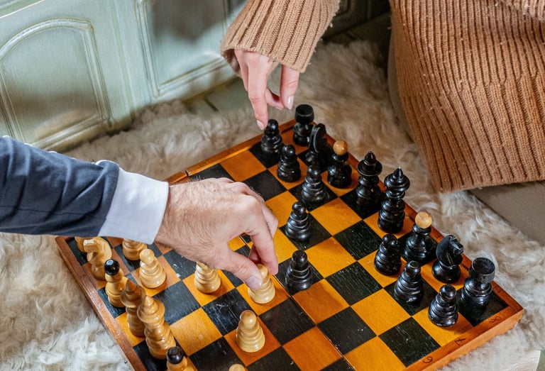 Two people playing a game of chess on a vintage wooden board over a soft white sheepskin rug.