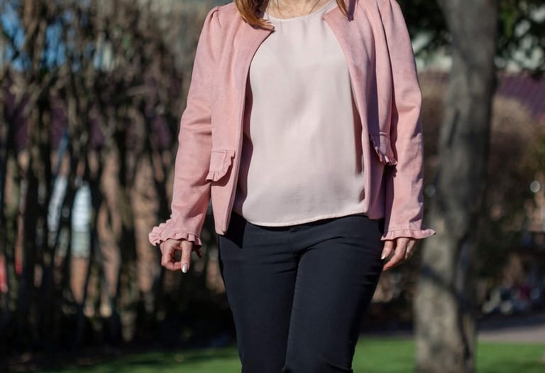 A woman wearing a stylish pink jacket and black trousers walking outdoors in a sunny park.