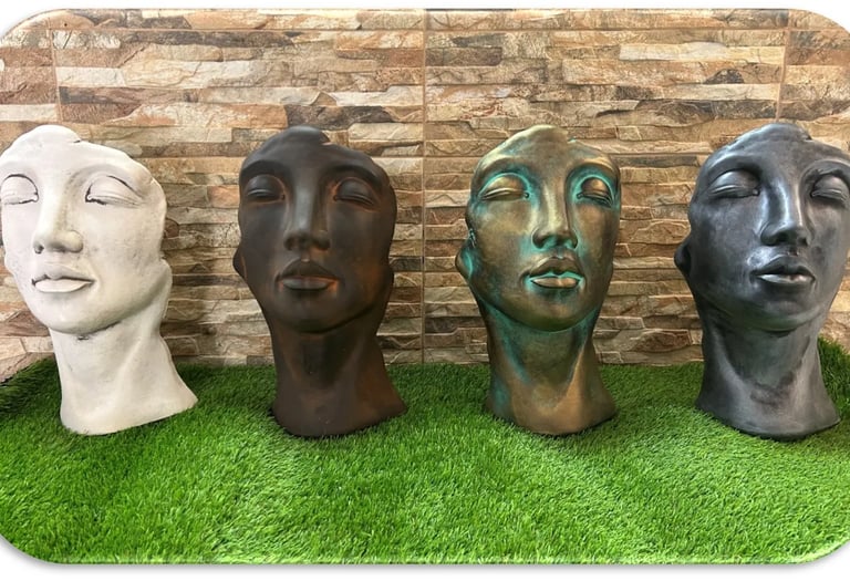 Four face-shaped garden planter pots in different finishes displayed on artificial grass.