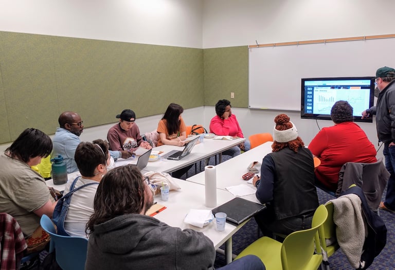 A group of people gathered around a table, looking intently at a presenter in the front of the room