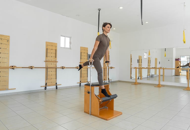Woman performing a standing leg exercise on a wooden Pilates Wunda Chair in a studio setting.