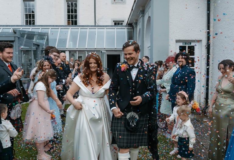 A smiling bride and groom in a traditional kilt walking through colorful wedding confetti outdoors.