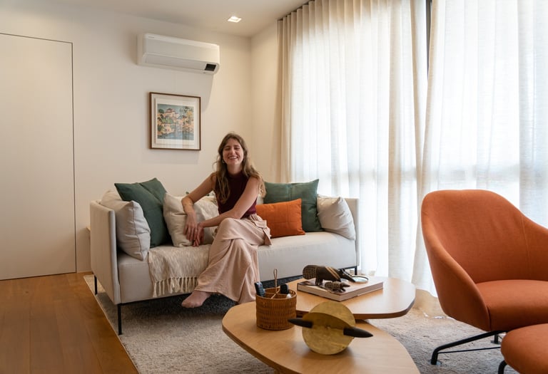 Mulher jovem sorrindo, sentada descalça em um sofá com almofadas coloridas, em uma sala aconchegante