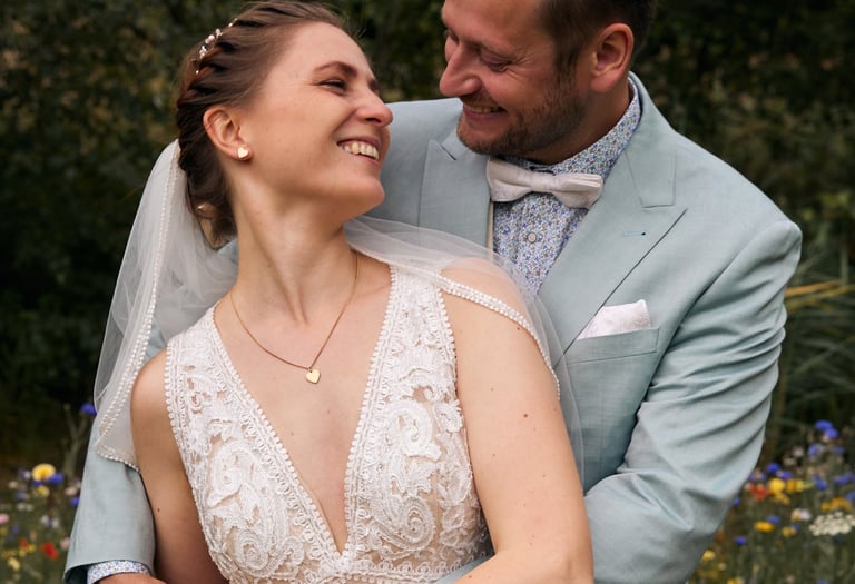 Smiling bride in lace wedding dress and groom in light blue suit posing in a wildflower garden.