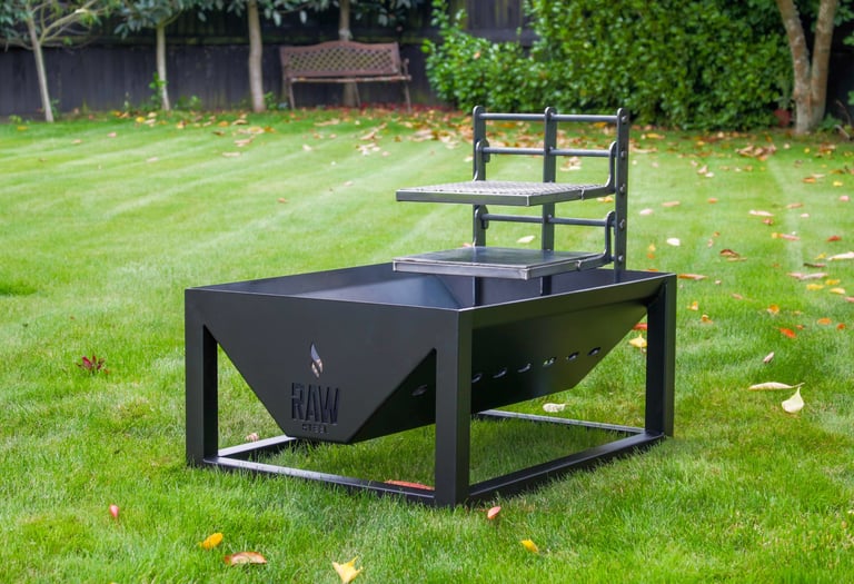 A black Raw Steel fire pit with an adjustable grill grate on a green backyard lawn.