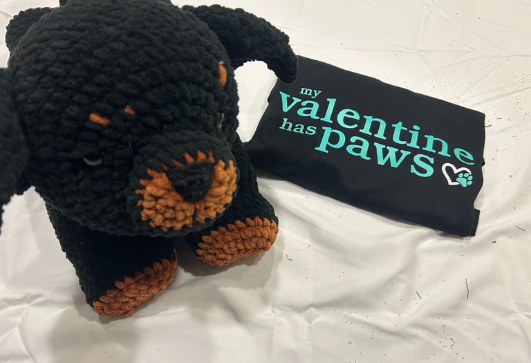 a folded tshirt that reads "my valentine has paws" sits beside a stuffed rottweiler