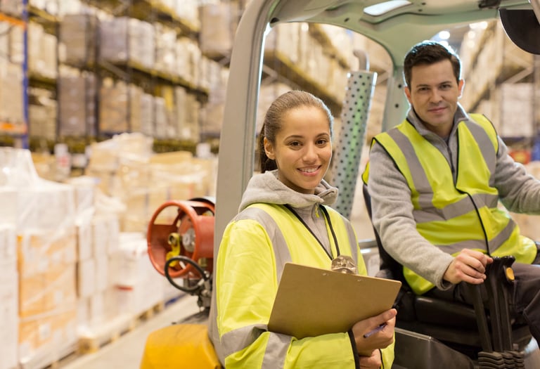 Trabajadores en un almacén usando chalecos reflectantes; uno manejando un montacargas y el otro con 