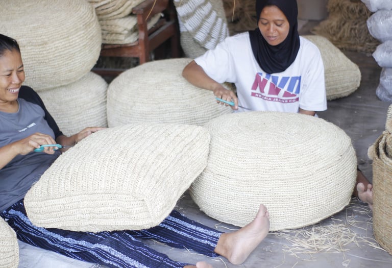 Two Indonesian artisans weaving and trimming handmade straw floor poufs and seagrass cushions.