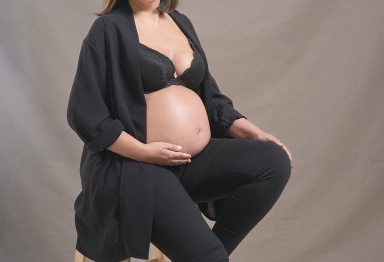 A pregnant woman in a black outfit poses for a maternity photoshoot sitting on a wooden stool.