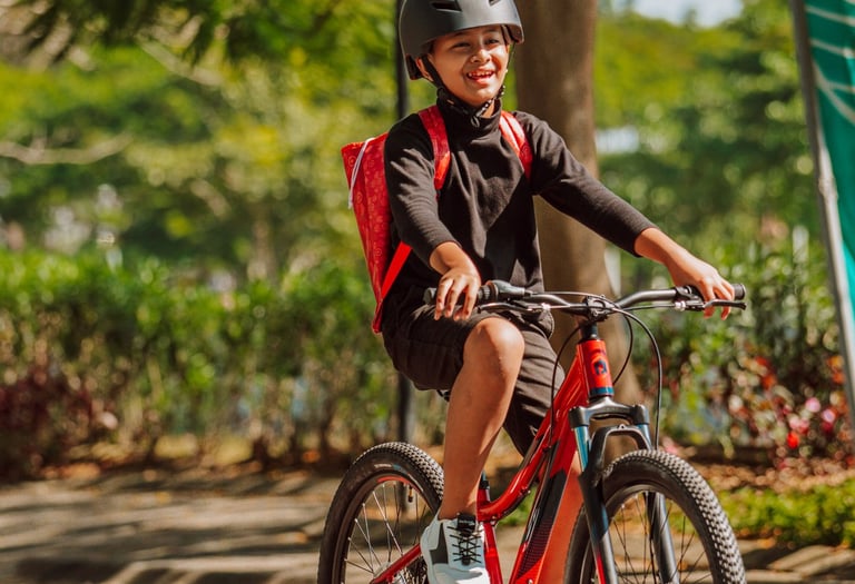 Smiling young boy wearing a helmet and backpack riding a red mountain bike on a sunny park path.