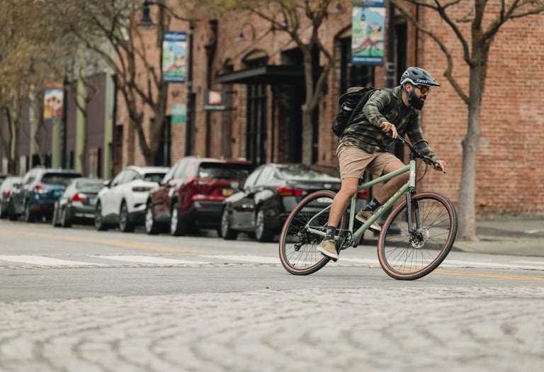 A male cyclist wearing a helmet and backpack rides a gravel bike through a city street.