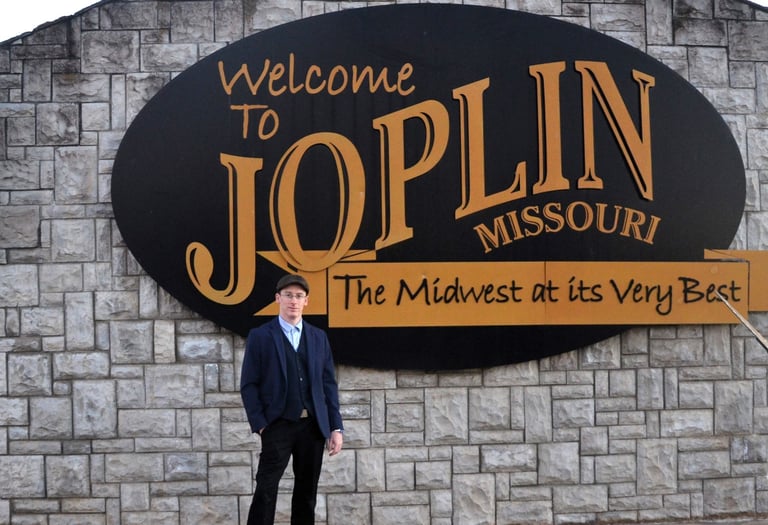 A man in a suit standing in front of the Welcome to Joplin Missouri sign on a stone wall.