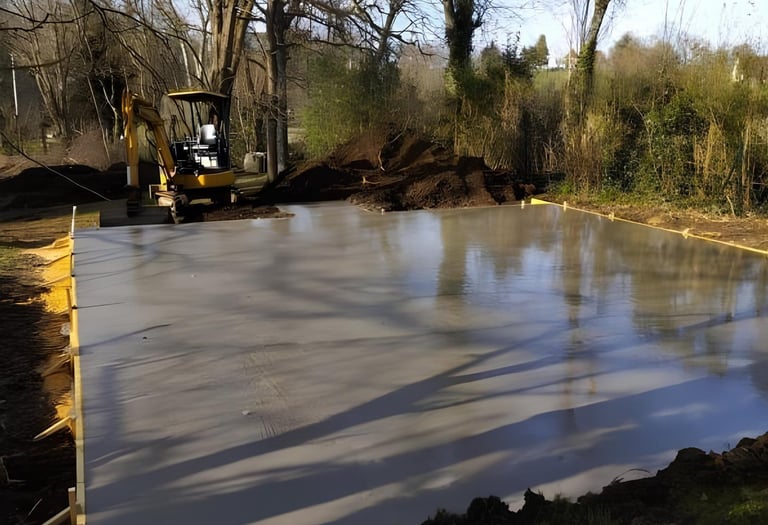 A freshly poured wet concrete slab foundation at a construction site with an excavator in the background.