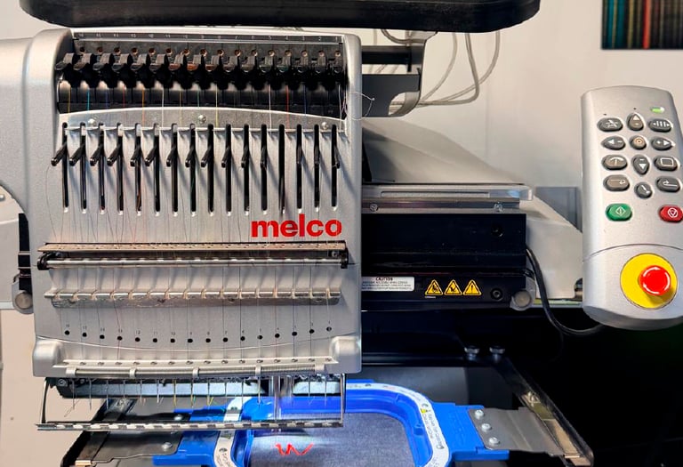 A multi-needle Melco commercial embroidery machine stitching a red logo onto a gray garment.